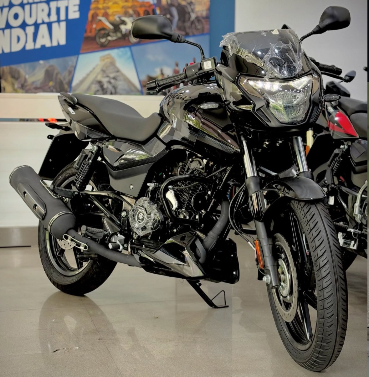 Black Bajaj Pulsar naked street bike parked inside an Indian showroom with plastic still on the headlamp cowl, front three-quarter view